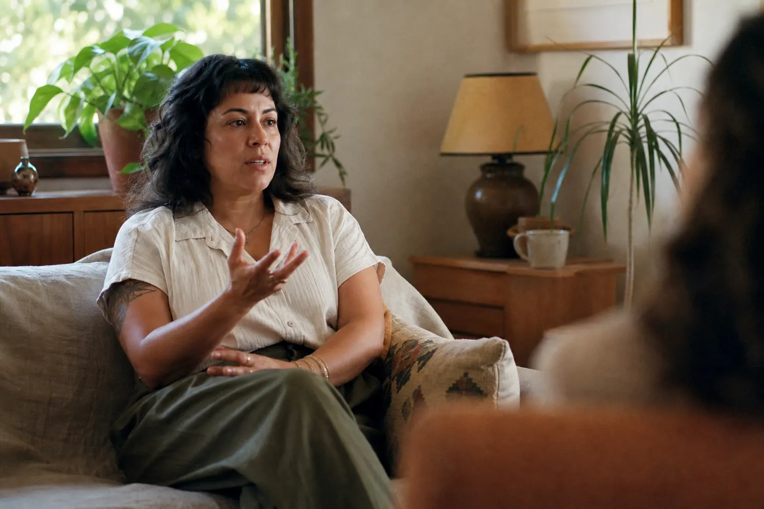 A woman in her thirties sitting on a couch in a sunlit therapy office, talking with her hands
