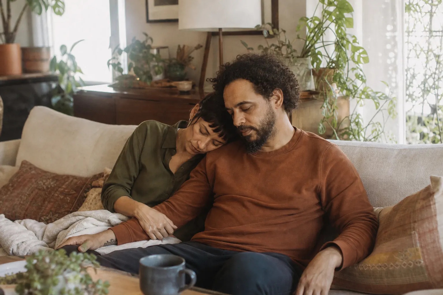 A couple in their thirties sitting close on a living room couch, one resting a hand on the other's knee